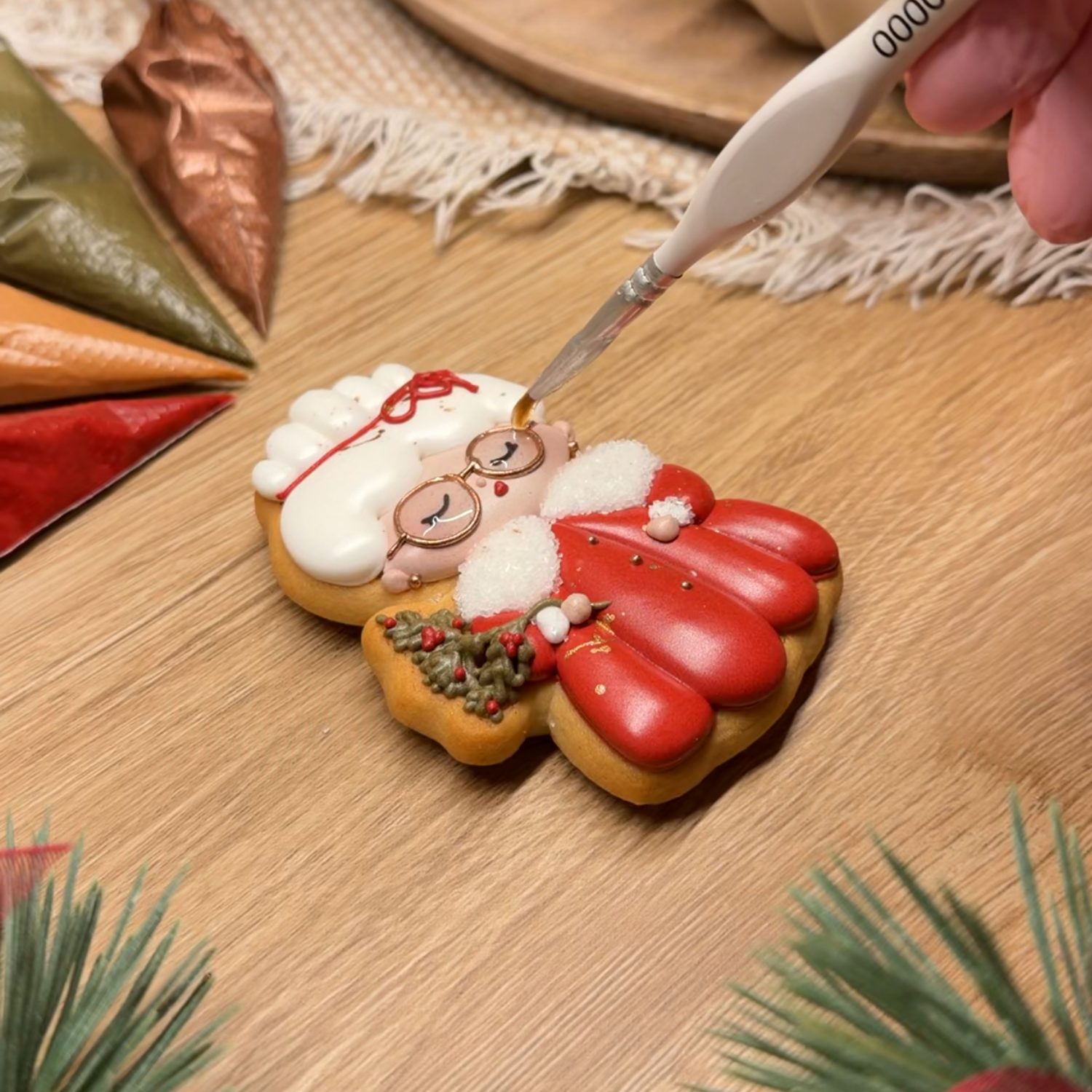 A fine brush adding gold glasses to a Mrs Claus cookie decorated with royal icing, next to piping bags of colored icing